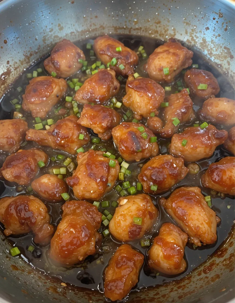 A skillet with golden-brown crispy chicken pieces sizzling in a rich, sticky Mongolian sauce, with fresh green onions sprinkled on top.